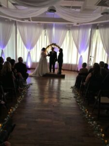 Couple in low light in front of wedding arch saying their vows with full length windows in the background covered by white drapes, 660 LaFayette Street, Horton Events Downtown Nashville Couple in low light in front of wedding arch saying their vows with full length windows in the background covered by white drapes, 660 LaFayette Street, Horton Events Downtown Nashville